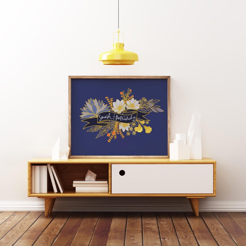 May include: A framed art print with a navy blue background features a floral design and the text "Smash Patriarchy" on a banner. The artwork includes white, yellow, and gold floral elements. The print is displayed on a wooden cabinet with a white drawer.
