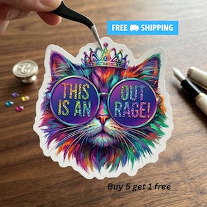 May include: A die-cut sticker featuring a colorful, multi-hued cat wearing a jeweled crown and large round glasses. The text "THIS IS AN OUTRAGE!" is printed in glittery letters across the lenses of the glasses. The sticker has a white border.