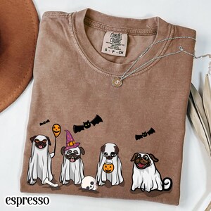 May include: A brown t-shirt featuring a graphic of four pugs dressed in white ghost costumes for Halloween. The pugs are accompanied by bats, a pumpkin, and a skull. The shirt has a Comfort Colors label at the neck and a silver-toned necklace.