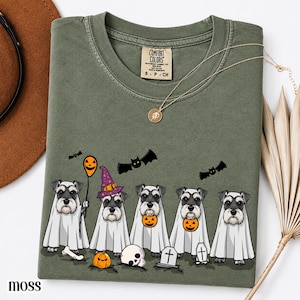 Puede incluir: Una camiseta verde oliva con una etiqueta Comfort Colors en el cuello. El gráfico muestra cinco perros Schnauzer disfrazados de fantasmas, uno de ellos con un sombrero de bruja morado. El diseño incluye calabazas naranjas, murciélagos, una calavera y lápidas.
