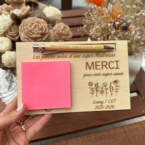 Peut inclure: Un support à notes rectangulaire en bois avec un stylo en bambou gravé 'Merci Maitresse - Lenny CE1 2025-2026'. La plaque porte les inscriptions 'Les petites notes d'une super Maîtresse', 'MERCI pour cette super année', 'Lenny / CE1 2025-2026', des motifs floraux et un bloc-notes adhésif rose vif.