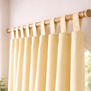 May include: Cream-colored curtains hanging from a wooden rod. The curtains are made of a soft fabric and are attached to the rod with fabric loops. The curtains are hanging in front of a window, and the light is coming through the window.