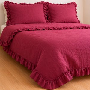 May include: A bed set featuring a deep red comforter and two matching pillow shams. The bedding has a ruffled edge detail, adding a decorative touch. The set is displayed on a bed with a light-colored mattress.