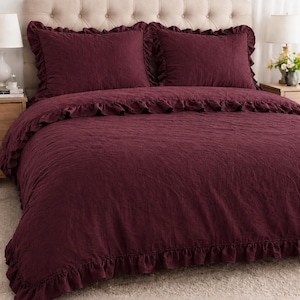 May include: A burgundy bedding set with ruffled edges. The set includes two pillow shams and a duvet cover, all in a solid color. The bed is made up with the bedding set, and the headboard is a neutral color.