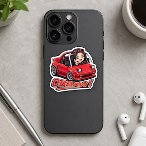 May include: A matte black smartphone featuring a sticker of a red sports car with pop-up headlights. An anime-style character sits in the driver seat making a peace sign. The text OBBAW! is printed in bold red and white letters below the car.