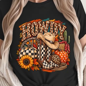 Puede incluir: Una camiseta negra con un estampado gráfico que muestra la palabra HOWDY en letras con estampado de leopardo. El diseño incluye un sombrero de vaquero, botas a cuadros, un girasol, calabazas, un cactus, una rueda de carreta y hojas de otoño.