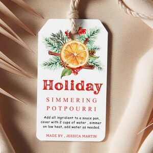 May include: A rectangular white gift tag with a top hole and string, featuring a watercolor illustration of an orange slice, pine needles, cinnamon sticks, and red berries. The text reads: Holiday SIMMERING POTPOURRI. Add all ingredient to a souce pan, cover with 2 cups of water , simmer on low heat, add water as needed. MADE BY , JESSICA MARTIN.