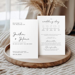 Op de afbeelding: Twee witte trouwuitnodigingskaarten met zwarte tekst. De linkerkaart zegt "Welkom bij de bruiloft van Jonathan + Juliana" en de rechterkaart heeft een trouwplanning. De kaarten liggen op een houten dienblad.