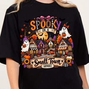 May include: A black t-shirt featuring a colorful graphic of a small town scene with ghosts, pumpkins, sunflowers, and an orange truck. The text reads "SPOOKY IN A Small Town" with the number "689883" displayed on a wooden sign at the bottom.