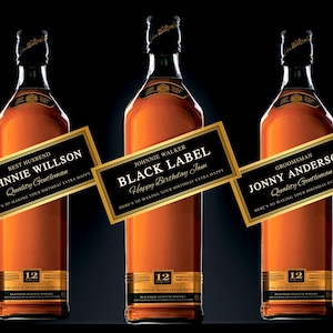 Op de afbeelding: Drie amberkleurige glazen flessen Johnnie Walker Black Label 12 jaar oude blended Scotch whisky. Elke fles heeft een aangepast rechthoekig zwart en goudkleurig etiket met tekst zoals Best Husband Johnnie Willson, Black Label Happy Birthday Jam en Groomsman Jonny Anderson, allemaal met de zin Here's to making your birthday extra happy.