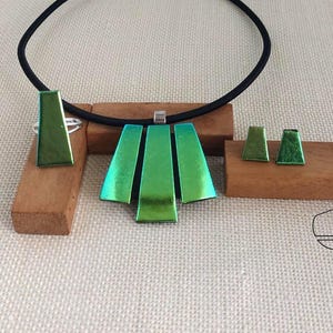 Op de afbeelding: Een moderne sieradenset met een groene hangerketting, bijpassende ring en oorbellen. De hanger heeft drie verticale groene panelen en de ring en oorbellen hebben een vergelijkbare groene kleur. De ketting heeft een zwart koord.