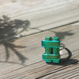 Op de afbeelding: Een ring met een rechthoekig, rastervormig sieraad van iriserend groen dichroïsch glas. Het glas heeft een getextureerd, glinsterend oppervlak met gouden en blauwe accenten. Het sieraad is bevestigd op een eenvoudige, zilverkleurige metalen ringband.