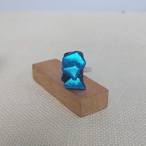 Op de afbeelding: Een ring met een centraal punt bestaande uit gelaagde, iriserende blauwe en groenblauwe glasstukjes met een metaalachtige glans. De glaselementen zijn op een donkere basis geplaatst en de ringband lijkt gemaakt van zilverkleurig metaal.