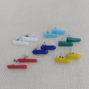 Op de afbeelding: Een verzameling kleurrijke oorstekers in verschillende tinten, waaronder rood, geel, blauw, groen en wit. Elke oorbel heeft een rechthoekig ontwerp met een afgerond uiteinde en een zilveren pin voor doorboorde oren.