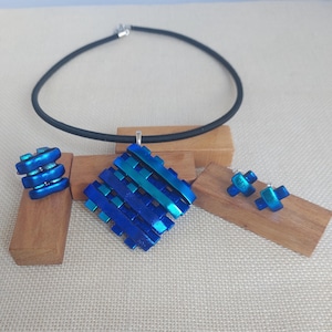 Op de afbeelding: Een moderne sieradenset met een blauwe hangerketting, bijpassende oorbellen en een ring. De hanger en ring hebben een geweven ontwerp en de oorbellen zijn kleine kruisjes. De ketting heeft een zwart koord.
