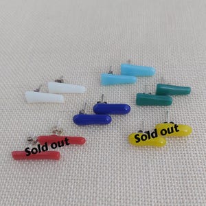 Op de afbeelding: Een verzameling kleurrijke oorstekers in verschillende vormen en kleuren, waaronder wit, blauw, groen, rood en geel. Sommige oorbellen hebben de tekst "Sold out". De oorbellen worden gepresenteerd op een neutrale achtergrond.