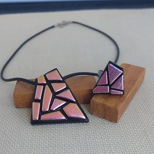 Op de afbeelding: Een sieradenset bestaande uit een hanger aan een zwart koord en een bijpassende ring. Beide stukken hebben een zwarte basis met een geometrisch mozaïekpatroon van iriserende, metallic roze scherven die het licht reflecteren met een glans.