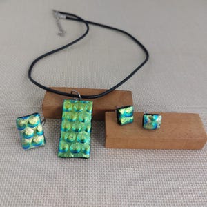 Op de afbeelding: Een sieradenset van dichroïsch glas met een ring, een hangerketting en oorstekers. De set heeft een getextureerd, bubbelachtig patroon in groen- en blauwtinten. De hanger hangt aan een zwart koord.
