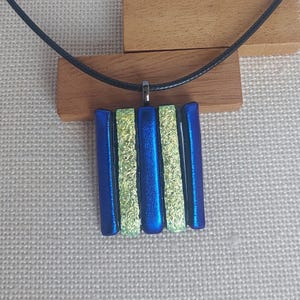 Puede incluir: Un collar con colgante azul y dorado. El colgante presenta barras rectangulares verticales de vidrio azul y dorado. El collar tiene un cordón negro y un cierre plateado. El colgante se muestra sobre una superficie de madera.