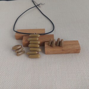 Op de afbeelding: Een sieradenset met een ketting, ring en oorbellen. De ketting heeft een zwart koord en een hanger van gestapelde, olijfgroene, langwerpige kralen. De ring en oorbellen hebben ook dezelfde olijfgroene, langwerpige kralen. De set wordt gepresenteerd op kleine houten blokken.