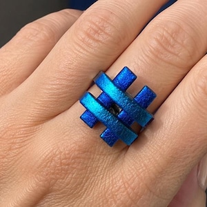 Op de afbeelding: Een ring met een geometrisch, rasterachtig ontwerp bestaande uit kruisende, getextureerde staven in levendige tinten metallic blauw. De structuur bestaat uit drie horizontale en drie verticale staven die een vierkant patroon vormen.