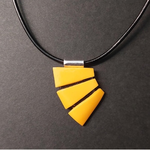 May include: A geometric pendant necklace featuring a black leather cord and a bright yellow pendant. The pendant is composed of three angled, rectangular shapes with black lines separating them. A silver-colored metal tube connects the pendant to the cord.