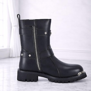 May include: Black leather ankle boot with a silver zipper, straps, and diamond-shaped studs. The boot has a chunky sole and a metal accent on the toe. The text "The Ultimate Leather" is at the bottom.