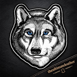May include: A black and white embroidered patch featuring the detailed head of a wolf with striking bright blue eyes. The design includes a thick white border and the text theultimateleather printed in the bottom right corner of the image.