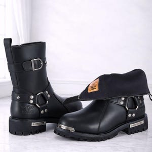 May include: Black leather motorcycle boots with silver-tone hardware, including buckles, rings, and studs. One boot is upright, the other has a folded-down cuff. The boots have a thick sole and a small metal plate with text. The Ultimate Leather is written at the bottom.