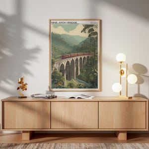 May include: A framed print of the Nine Arch Bridge in Sri Lanka, with a red train crossing the bridge. The artwork depicts green hills and a light blue sky. The print is displayed on a light wood cabinet, with a gold lamp.