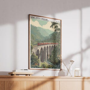 May include: A framed print of the Nine Arch Bridge in Sri Lanka. The image shows a red train on the bridge, surrounded by green hills and trees. The print has a vintage style, reminiscent of travel posters.