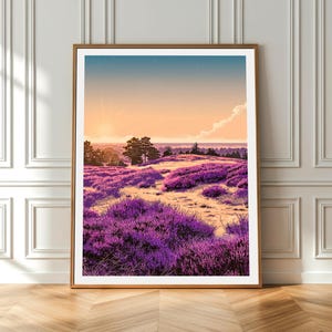 May include: A framed landscape art print featuring a field of purple flowers under a sunset sky. The artwork has a warm color palette with shades of purple, orange, and blue. The print is displayed on a wooden floor against a white wall.