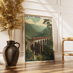 May include: A framed print of the Nine Arch Bridge in Sri Lanka. The artwork shows a red train crossing the bridge, surrounded by green mountains and trees. The print is in a gold frame and leans against a white wall.