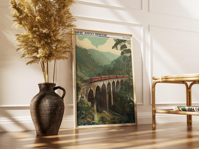 May include: A framed print of the Nine Arch Bridge in Sri Lanka. The artwork shows a red train crossing the bridge, surrounded by green mountains and trees. The print is in a gold frame and leans against a white wall.