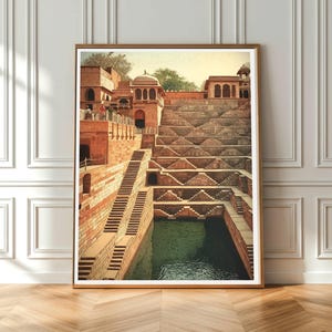 May include: Framed print of a stepwell with intricate architecture. The image showcases a series of descending steps leading to a water reservoir. The structure is made of reddish-brown stone, with a green-tinted water at the bottom. The print is displayed in a wooden frame.