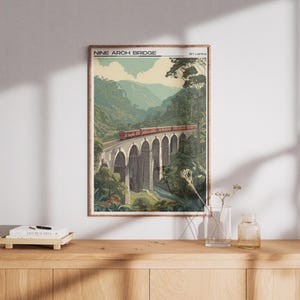 May include: A framed print of the Nine Arch Bridge in Sri Lanka. The artwork shows a red train crossing the bridge, surrounded by green foliage and mountains. The print is displayed on a wooden surface.