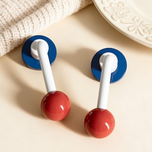 May include: Two decorative cabinet knobs. Each knob features a red sphere, a white stem, and a blue circular base. The knobs are designed with a modern aesthetic and are likely used for drawers or cabinets.
