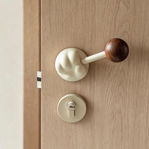 May include: A close-up of a door handle and lock set. The handle features a white, organic-shaped base with a white arm and a dark brown wooden ball. The lock and base are a matching off-white color, set against a light wood door.