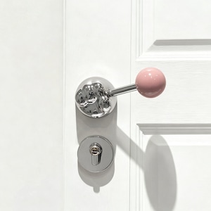 May include: A chrome door handle with a fist-shaped backplate and a pink spherical handle. The door hardware is mounted on a white door, with a matching chrome keyhole below the handle.