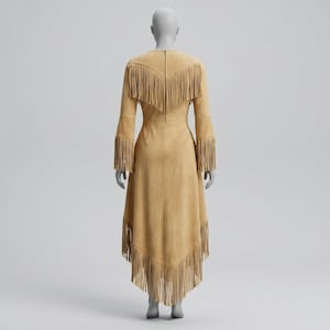 May include: A tan suede dress featuring long sleeves and a high-low hemline. The garment is adorned with layers of fringe detailing across the shoulders, back, and hem. A vertical zipper closure is visible along the center of the back panel.