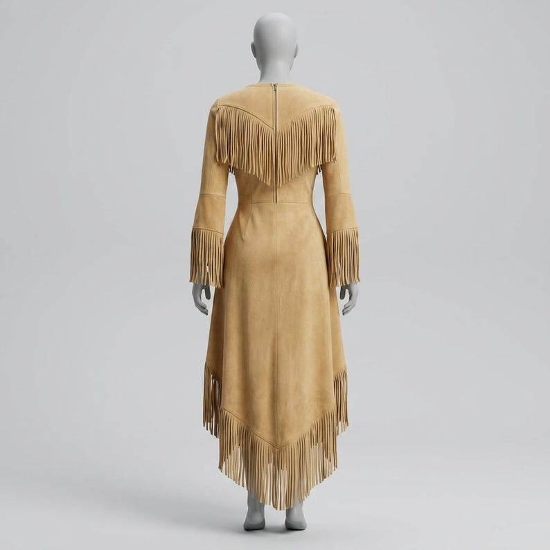 May include: A tan suede dress featuring long sleeves and a high-low hemline. The garment is adorned with layers of fringe detailing across the shoulders, back, and hem. A vertical zipper closure is visible along the center of the back panel.