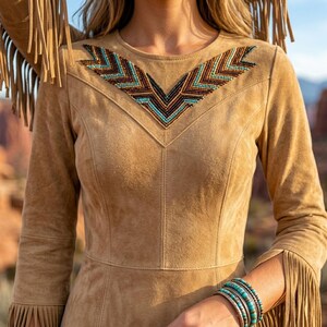 May include: A tan suede long-sleeve dress featuring a V-shaped beaded pattern across the chest in shades of turquoise, brown, and black. The garment includes long fringe detailing along the sleeves and shoulders, and a fitted bodice design.