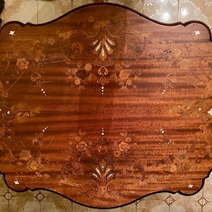 May include: An oval wooden table with a scalloped edge featuring intricate floral marquetry and inlaid mother-of-pearl accents. The surface displays a rich reddish-brown wood grain with symmetrical botanical patterns and decorative scrollwork.