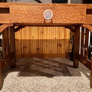 May include: A medium-brown oak wood desk featuring a rectangular top, four sturdy legs, and a single central drawer. The drawer is equipped with a round, light-colored knob. The desk displays a prominent wood grain pattern and decorative corbels.
