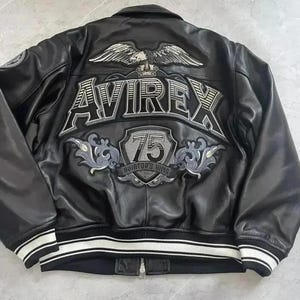 Puede incluir: Una chaqueta de cuero negra con un águila grande bordada, el texto AVIREX y un emblema de escudo con el número 75 y las palabras AVIATOR'S KING en la espalda. La chaqueta tiene un dobladillo y puños acanalados con rayas blancas y negras.