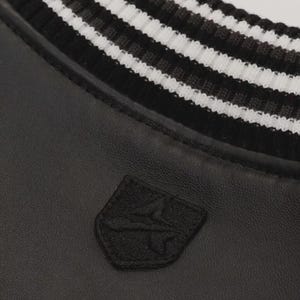 May include: Close-up of a black jacket with a white and black striped collar. A small, black embroidered patch is visible on the jacket's front. The jacket appears to be made of leather or a similar material.