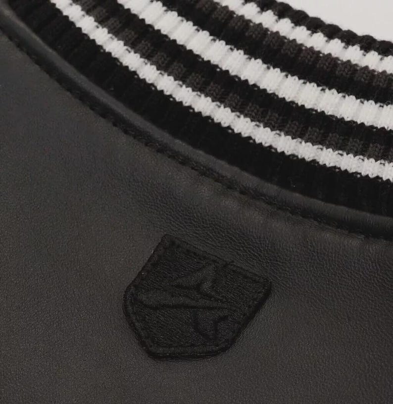 May include: Close-up of a black jacket with a white and black striped collar. A small, black embroidered patch is visible on the jacket's front. The jacket appears to be made of leather or a similar material.