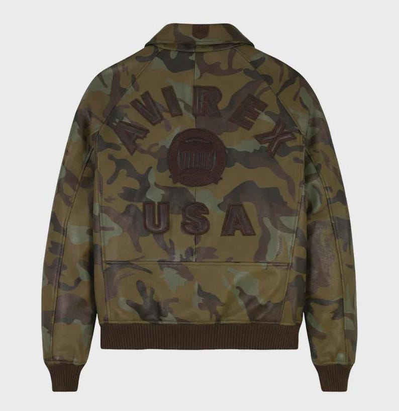 May include: A camouflage-patterned leather jacket with the word "AVIREX" arched across the back, along with a circular emblem and the word "USA" below. The jacket features a brown collar, cuffs, and hem.