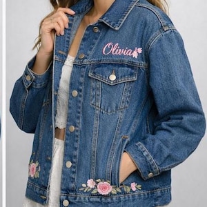 May include: A blue denim jacket with the name "Olivia" embroidered in pink script on the chest. The jacket features floral embroidery in pink and green at the hem. It has a classic collar, button closures, and pockets.