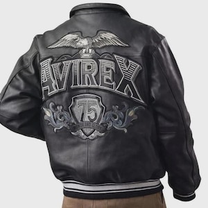 Puede incluir: Chaqueta de cuero negra con cuello y puños a rayas blancas y grises. La parte trasera presenta un gran diseño bordado con un águila, la palabra "AVIREX" y el texto "75 HONOR AND GLORY".